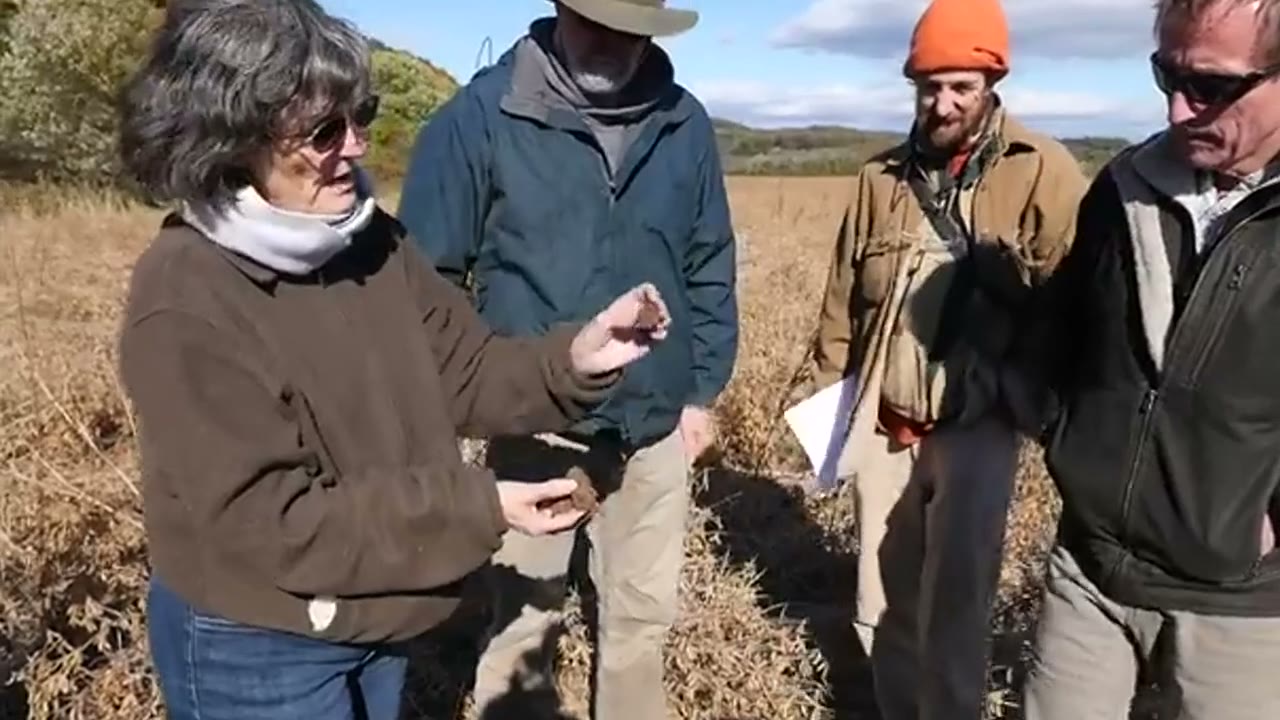 Dr. Elaine Ingham teaching a small group in a golden autumn field, a clod of soil in her hand.